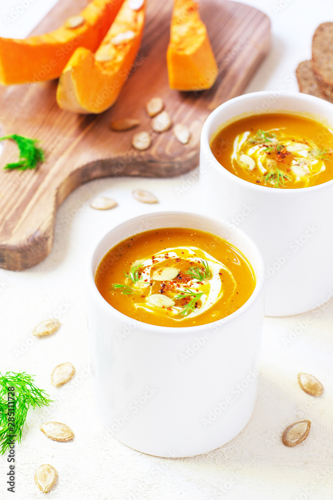 Roasted pumpkin and carrot soup with cream and pumpkin seeds on two white mug on white background with black bread slices.Vegetarian eating.Diet and healthy food concept.Space for copy and text Roasted pumpkin and carrot soup with cream and pumpkin seeds on two white mug on white background with black bread slices.Vegetarian eating.Diet and healthy food concept.Space for copy and text
