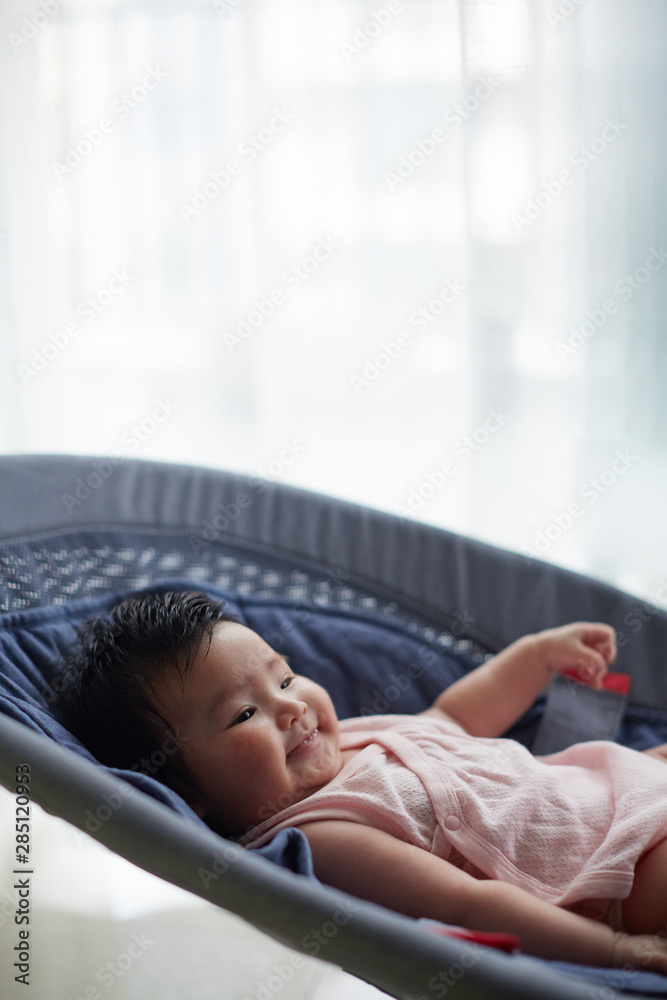 Cute Asian baby, sitting on a small lounger and playing with himself, at home