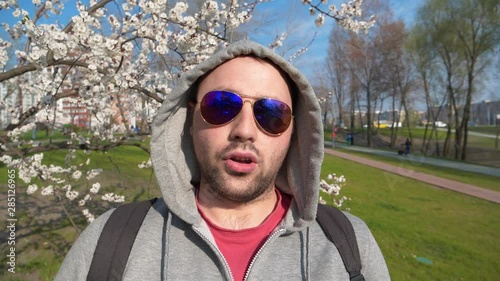 Man looks at the camera and speaks. Adult man stands near the apricot tree in city park and talks into the camera photo