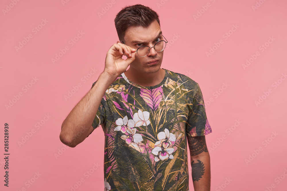 Portrait of young man in flowered t-shirt with confused face expression, looking through glasses with doubts, isolated over pink background Portrait of young man in flowered t-shirt with confused face expression, looking through glasses with doubts, isolated over pink background