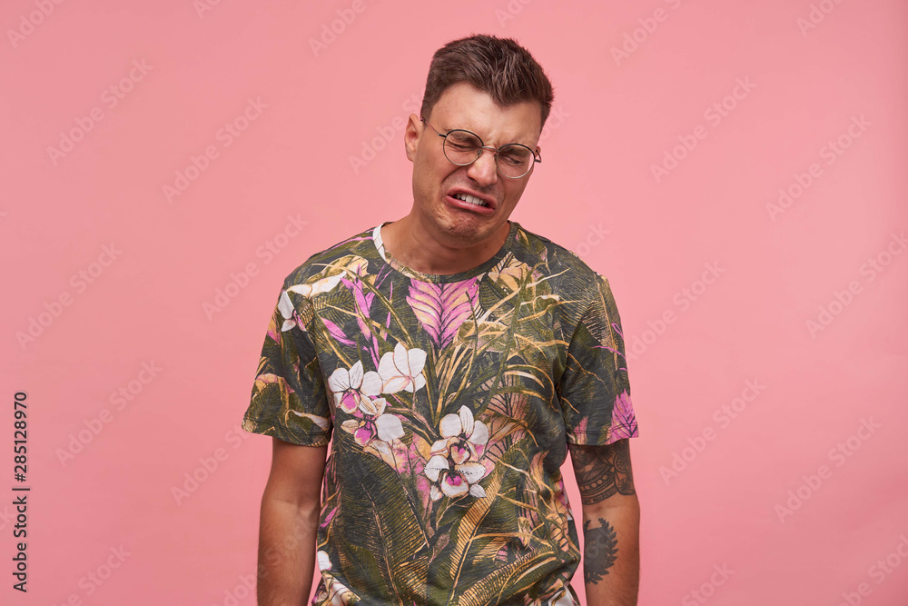 Shot of sad crying young man in glasses, wearing flowered t-shirt, looking unhappy and upset, standing over pink background, negative emotions concept Shot of sad crying young man in glasses, wearing flowered t-shirt, looking unhappy and upset, standing over pink background, negative emotions concept