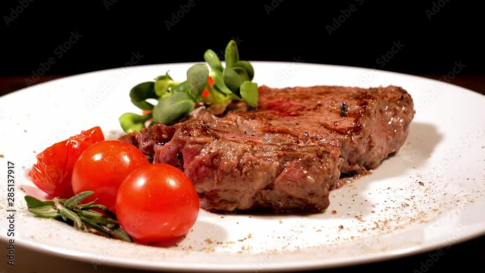 A piece of fried meat garnished with Cherry Tomatoes and microgreen rotating on a white plate. Tasty grilled steak close-up. Demonstration of the finished dish. A piece of fried meat garnished with Cherry Tomatoes and microgreen rotating on a white plate. Tasty grilled steak close-up. Demonstration of the finished dish.