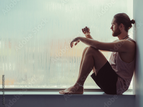 Side view of relaxed thoughtful male sitting in harmony on sill near against steamy window and looking down photo