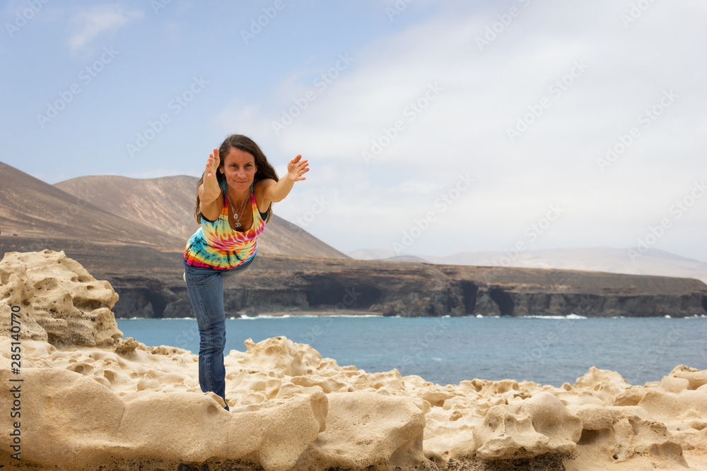 Middle aged woman in colorful urban outfit practices warrior yoga pose on rocks by the sea in Fuerteventura, Canary Islands. Female yogi in Virabhadrasana three on the coast Middle aged woman in colorful urban outfit practices warrior yoga pose on rocks by the sea in Fuerteventura, Canary Islands. Female yogi in Virabhadrasana three on the coast