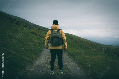 Back view of unrecognizable male with backpack standing on rough path on grassy hill slope against gray overcast sky in nature photo