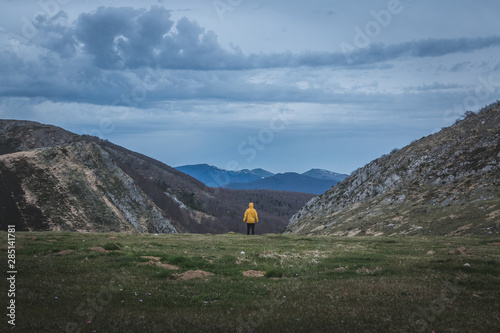 Back view of tourist in yellow coat looking at rough hills and overcast sky during trip in countryside photo