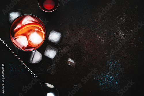 Negroni alcoholic cocktail beverage with dry gin, red vermouth and red bitter, bloody orange slice and ice cubes. Brown bar counter background, steel bar tools, top view, with copy space
