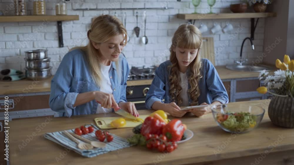 Mom and daughter cook together in a large bright kitchen. Mom cuts juicy peppers, a girl reads a recipe on a tablet 4K Mom and daughter cook together in a large bright kitchen. Mom cuts juicy peppers, a girl reads a recipe on a tablet 4K