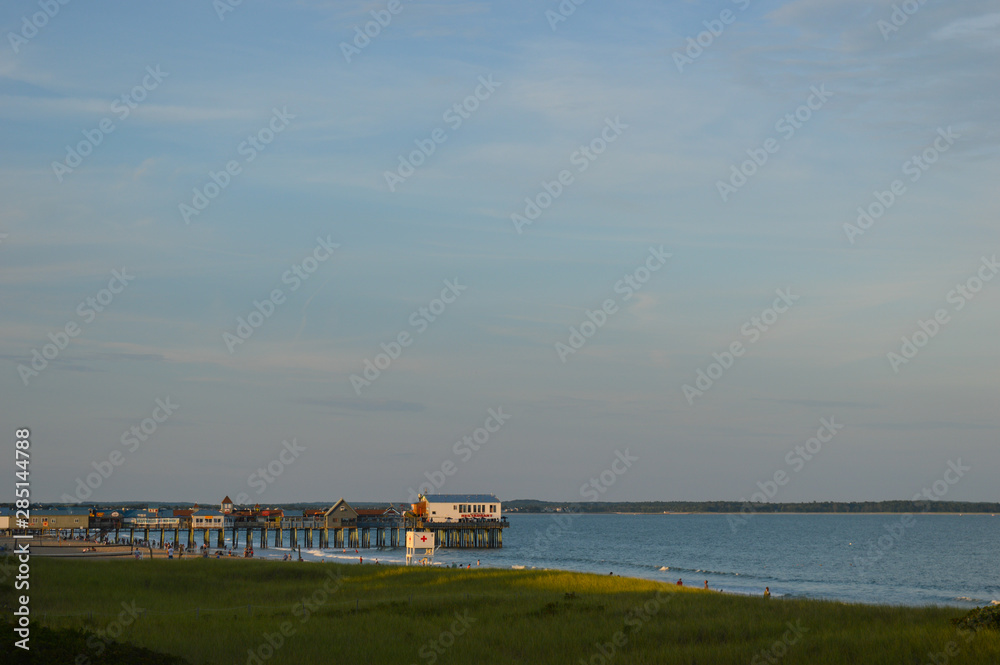 A pier lined with shopping and dining options extends from the Maine coast into the ocean. The sun is setting, bathing the scene in a golden glow.