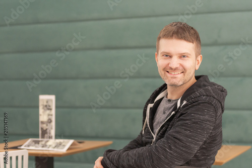 Photo shoot in cafe. Smiling openly at the camera. Man sitting, looking at the camera, right. On a chair, half turned.