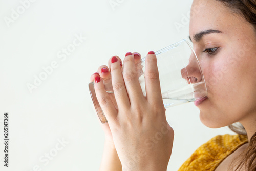 Close up of one beautiful girl with long hair and red-painted nails drinking fresh water in glass cup on light wall background. Health  healthy life  refresh and happiness concept.