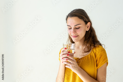 Close-up of one beautiful girl with smile, long hair and red-painted nails holding glass cup with water on light wall background. Health, healthy life, refresh and happiness concept.
