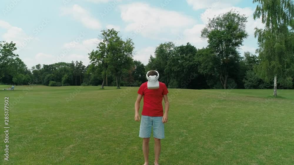 A man adjusted his VR headset and looking down in the park. Young guy in helmet experiencing augmented reality on the background of nature. 4K drone footage. A man adjusted his VR headset and looking down in the park. Young guy in helmet experiencing augmented reality on the background of nature. 4K drone footage.