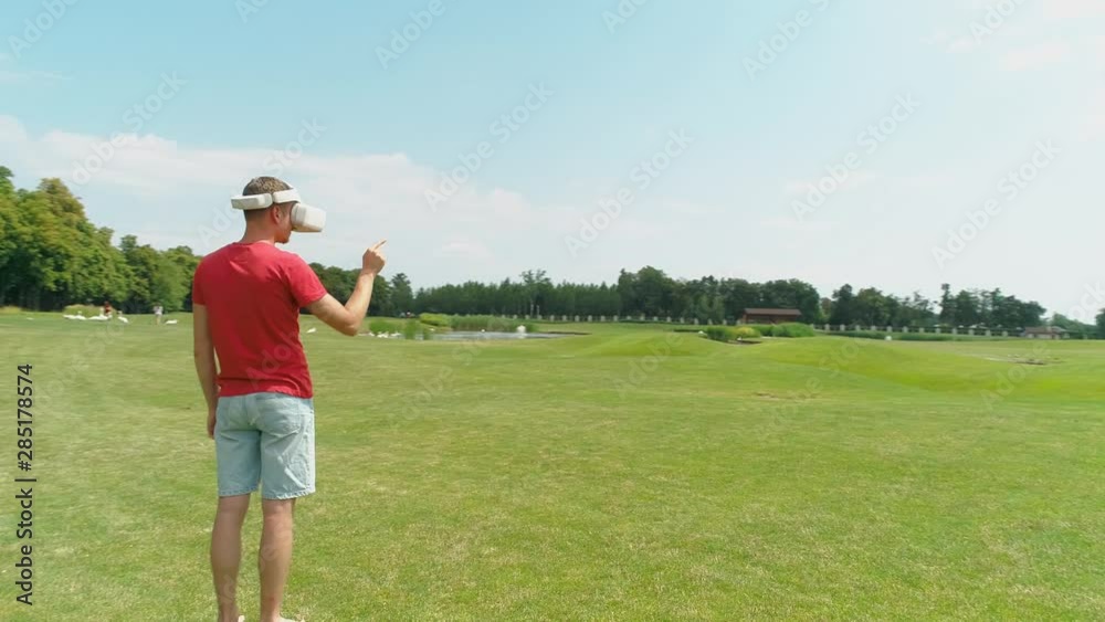 A man with virtual reality headset on his head is standing and using VR helmet on green grass among beautiful forest, blue sky with clouds on sunny day in summer time. 4K drone footage. A man with virtual reality headset on his head is standing and using VR helmet on green grass among beautiful forest, blue sky with clouds on sunny day in summer time. 4K drone footage.
