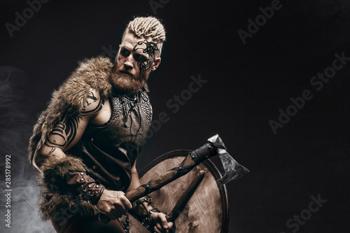 Vászonkép Medieval warrior berserk Viking with tattoo on skin, red beard and braids in hair with axe and shield attacks enemy