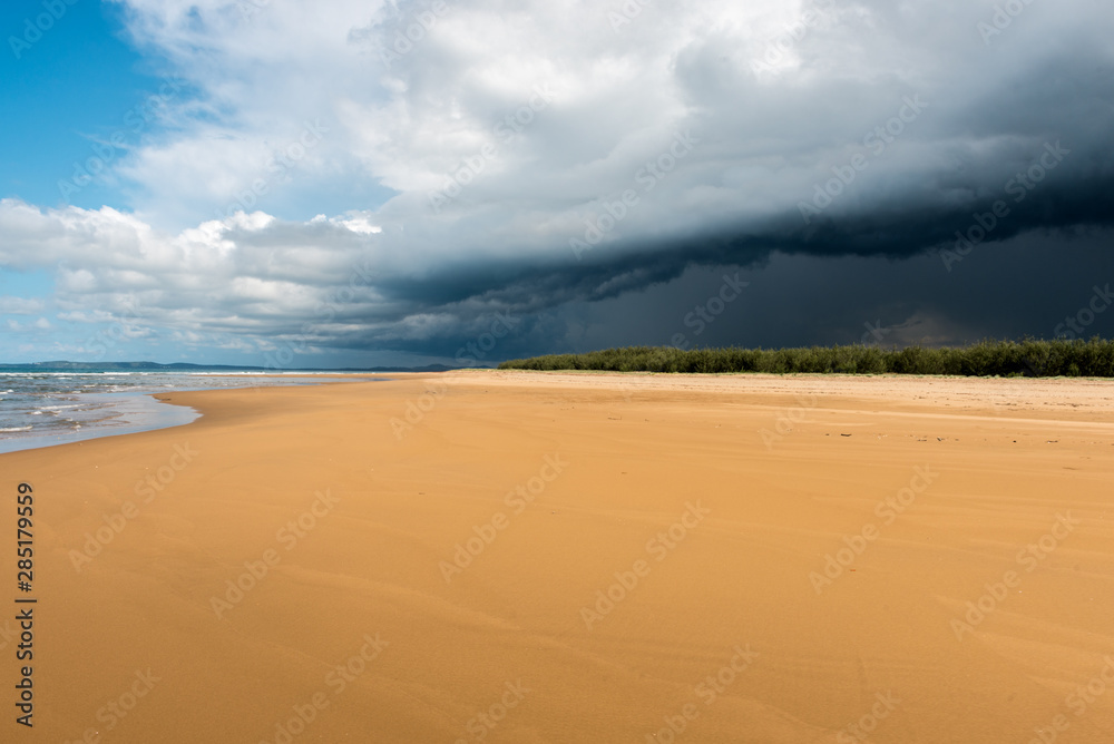 The sea water softs the bright orange sands as a thunderstorm bring dark dramatic clouds that loom over the lush and dense forests on Middle Island near 1770 in Australia.