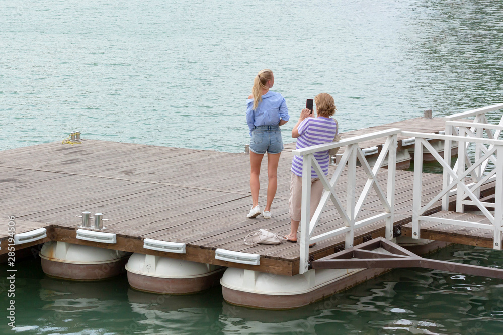 A woman aged photographs a young slender girl, her daughter, on the pier near the lake. A woman aged photographs a young slender girl, her daughter, on the pier near the lake.