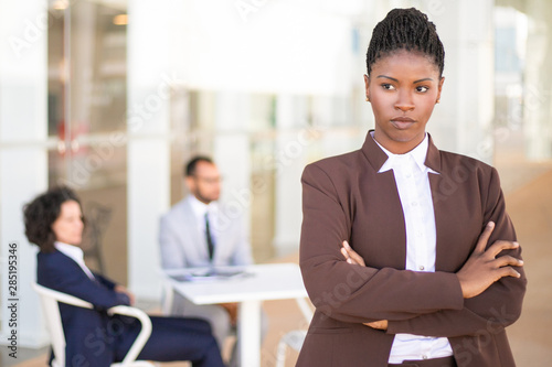 Stressed African American female employee standing with arms crossed and looking away. Her male and female colleagues sitting in background. Successful business woman concept