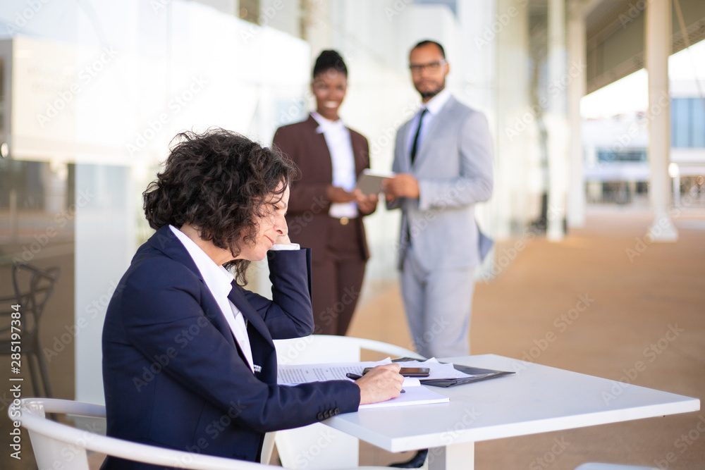 Tired unhappy female employee sitting in outdoor cafe and checking notes and papers. Her multiethnic colleagues standing in background and laughing. Office conflict concept Tired unhappy female employee sitting in outdoor cafe and checking notes and papers. Her multiethnic colleagues standing in background and laughing. Office conflict concept