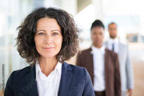 Successful female leader posing with her team in blurred background. Middle aged businesswoman smiling at camera, her two colleagues standing behind her. Successful team leader concept photo