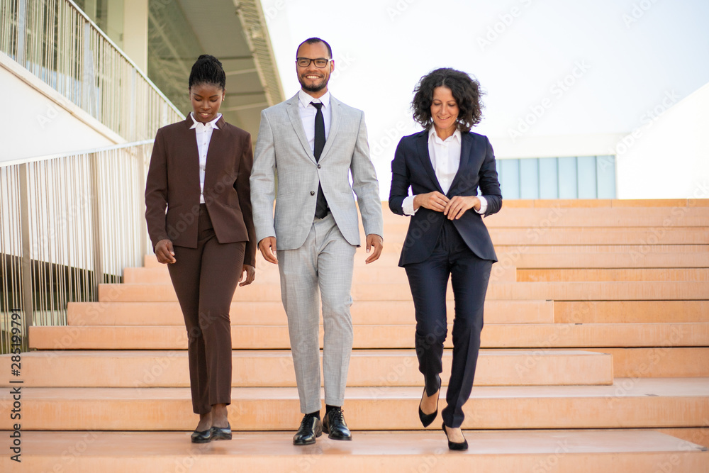 Happy business partners walking near office building. Multiethnic team of businesspeople going down stairs, talking, laughing. Successful teamwork or partnership concept Happy business partners walking near office building. Multiethnic team of businesspeople going down stairs, talking, laughing. Successful teamwork or partnership concept