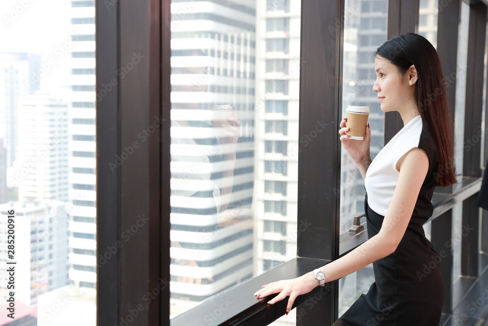 side view of asian businesswoman or lawyer in black and white dress with confident face who standing and holding cup of coffee in office and look away with city view background
