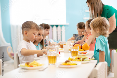 Kids have a dinner in kindergarten. Little boys and girls from the group of children sitting at table with lunch and eat appetizing. Children with caregiver in day care centre. Childs have healthy photo
