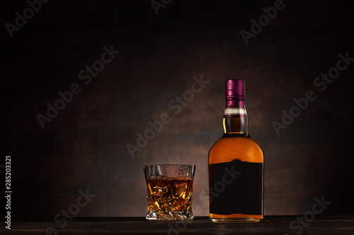 Glass of whiskey with ice and bottle on dark wood background