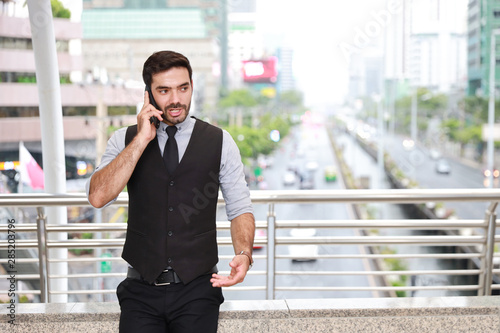 front view successful caucasian businessman in black suite who standing in downtown, he using cell phone with city background