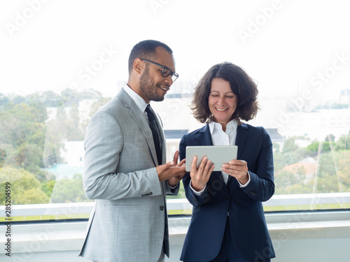 Two cheerful colleagues discussing team project during work break. Happy business man and woman standing indoors at office window, looking at tablet screen, talking and smiling. Teamwork concept