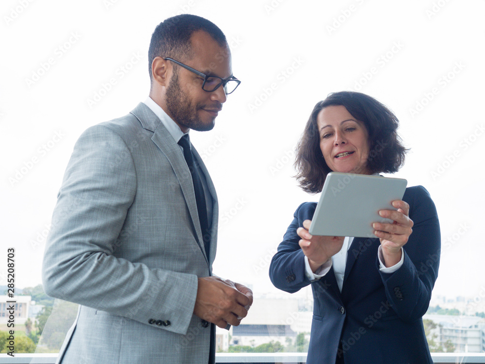 Satisfied business leaders watching project presentation on tablet. Business man and woman standing indoors at office window, woman holding tablet and talking. Business communication concept Satisfied business leaders watching project presentation on tablet. Business man and woman standing indoors at office window, woman holding tablet and talking. Business communication concept