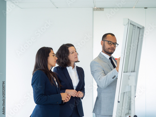 Serious male business leader explaining strategy to female colleagues. Business man drawing on board, his two coworkers looking at drawing. Presenting concept