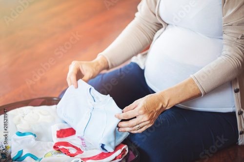 Pregnant woman packing suitcase, bag for maternity hospital at home, getting ready for newborn birth, labor. Pile of baby clothes, necessities and pregnant women at awaiting.