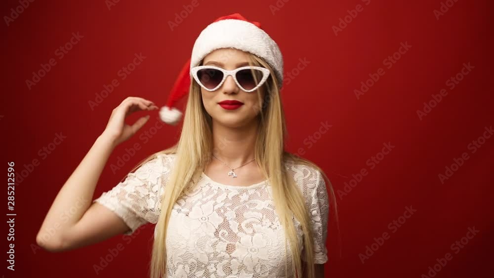 Happy charming blonde in sunglasses with santa claus hat. Red isolated background. Close-up portrait.