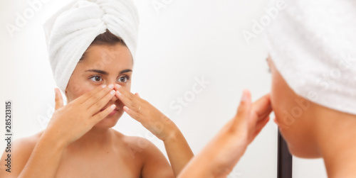 Portrait of a beautiful young woman applying face cream and looking at mirror in bathroom. Youthful girl with perfect skin using facial cosmetics. Skin care and health concept..