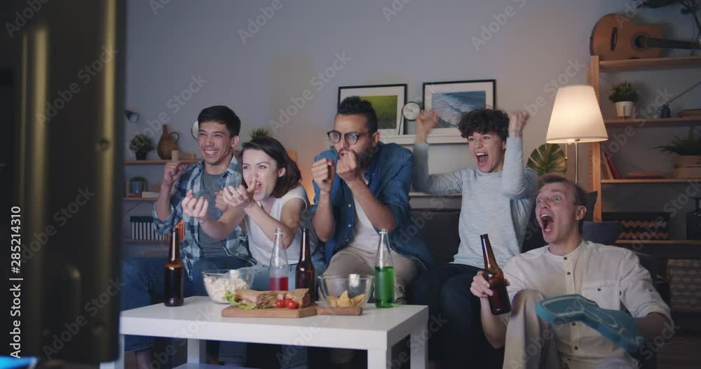 Slow motion of happy young people friends watching sports at night at home having fun doing high-five and laughing. Happiness and modern lifestyle concept. Slow motion of happy young people friends watching sports at night at home having fun doing high-five and laughing. Happiness and modern lifestyle concept.