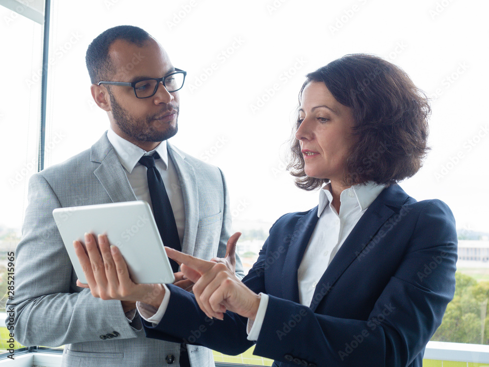 Serious female manager showing report on tablet to boss. Business man and woman standing indoors, using tablet and talking. Digital report concept Serious female manager showing report on tablet to boss. Business man and woman standing indoors, using tablet and talking. Digital report concept