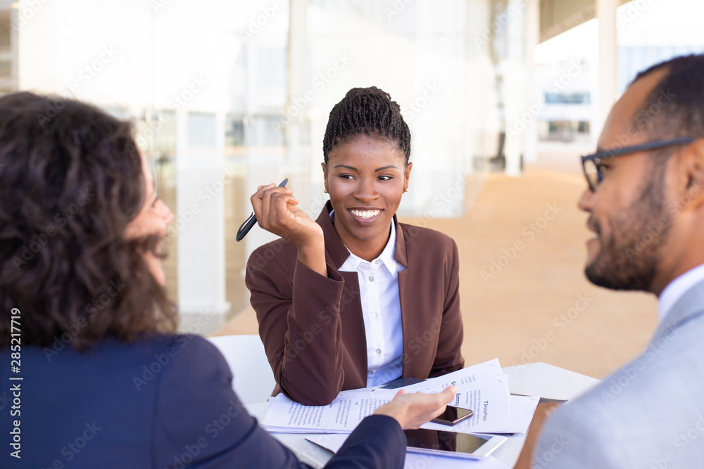 © Mangostar - Happy African American business woman discussing deal with partners. Multiethnic business partners sitting at table with documents in outdoor cafe and talking. Partnership concept