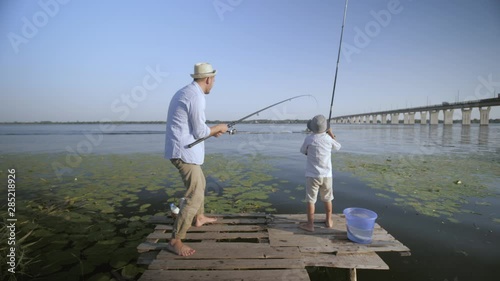 entertaining father and son, cheerful fisherm n with little boy in straw hats on pier near river catch fish while on vacation in village against a blue sky and water photo