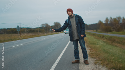 Casualy Dressed Hitchhiker Traveler Standing by the Highway and Thumbing at a Cloudy Day.