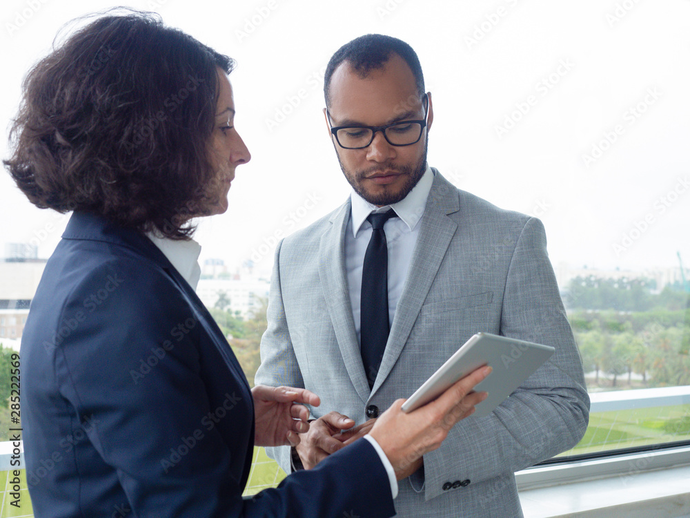 Serious business leaders watching content on tablet and discussing deal. Business man and woman standing indoors at office window, woman holding tablet and talking. Business communication concept Serious business leaders watching content on tablet and discussing deal. Business man and woman standing indoors at office window, woman holding tablet and talking. Business communication concept