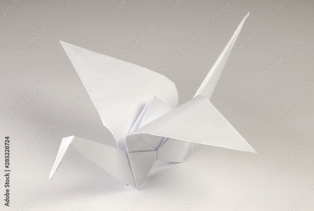 Light gray origami crane on gray background. Tsuru. Japanese art of paper folding. Flat square sheet of paper transferred into finished sculpture through folding and sculpting. Close up. Macro photo. Light gray origami crane on gray background. Tsuru. Japanese art of paper folding. Flat square sheet of paper transferred into finished sculpture through folding and sculpting. Close up. Macro photo.