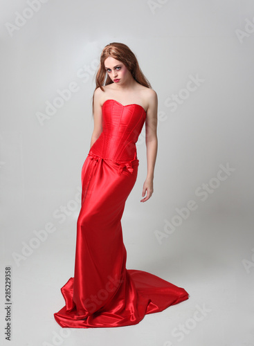 full length portrait of a  girl wearing a long red silk gown, Standing pose on a grey studio background.