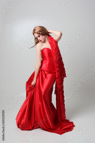 full length portrait of a girl wearing a long red silk gown, Standing pose on a grey studio background.