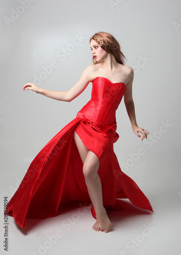 full length portrait of a girl wearing a long red silk gown, Standing pose on a grey studio background.