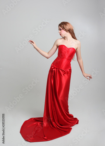 full length portrait of a girl wearing a long red silk gown, Standing pose on a grey studio background.