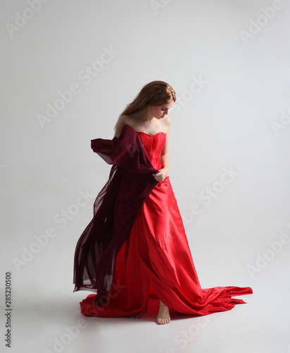 full length portrait of a  girl wearing a long red silk gown, Standing pose on a grey studio background.