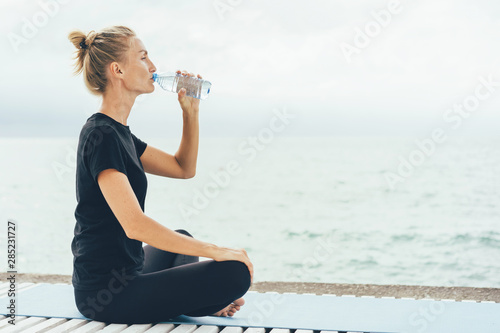 A woman during a sports training outside on the seashore drinks water to replenish the body's water balance. Sports and healthy concept. Yoga and meditation alone.
