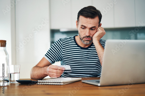 Sad man sitting at home with laptop and looking at credit card photo