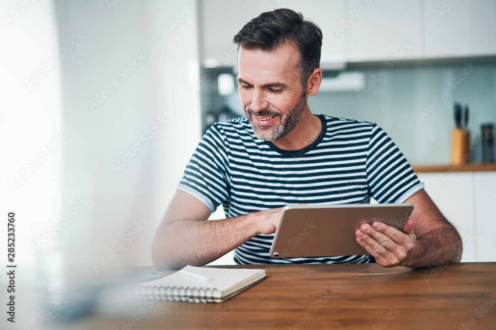 Smiling man sitting at home dining table and using tablet managing personal finances Smiling man sitting at home dining table and using tablet managing personal finances